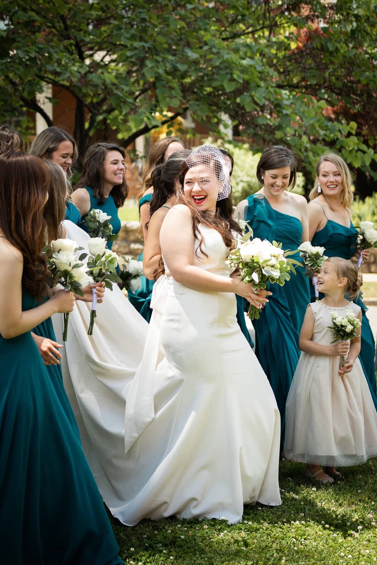  A joyful bride in a sleek satin wedding gown with a vintage birdcage veil laughs with her bridesmaids dressed in deep teal dresses. The group is captured mid-motion, holding bouquets of white roses, as they celebrate outdoors in a lush, green garden in Tennessee