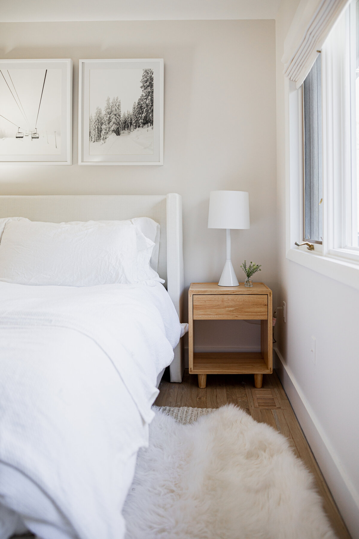 Bright bedroom with a white bed, a bedside table with a lamp, and a large window with natural light.