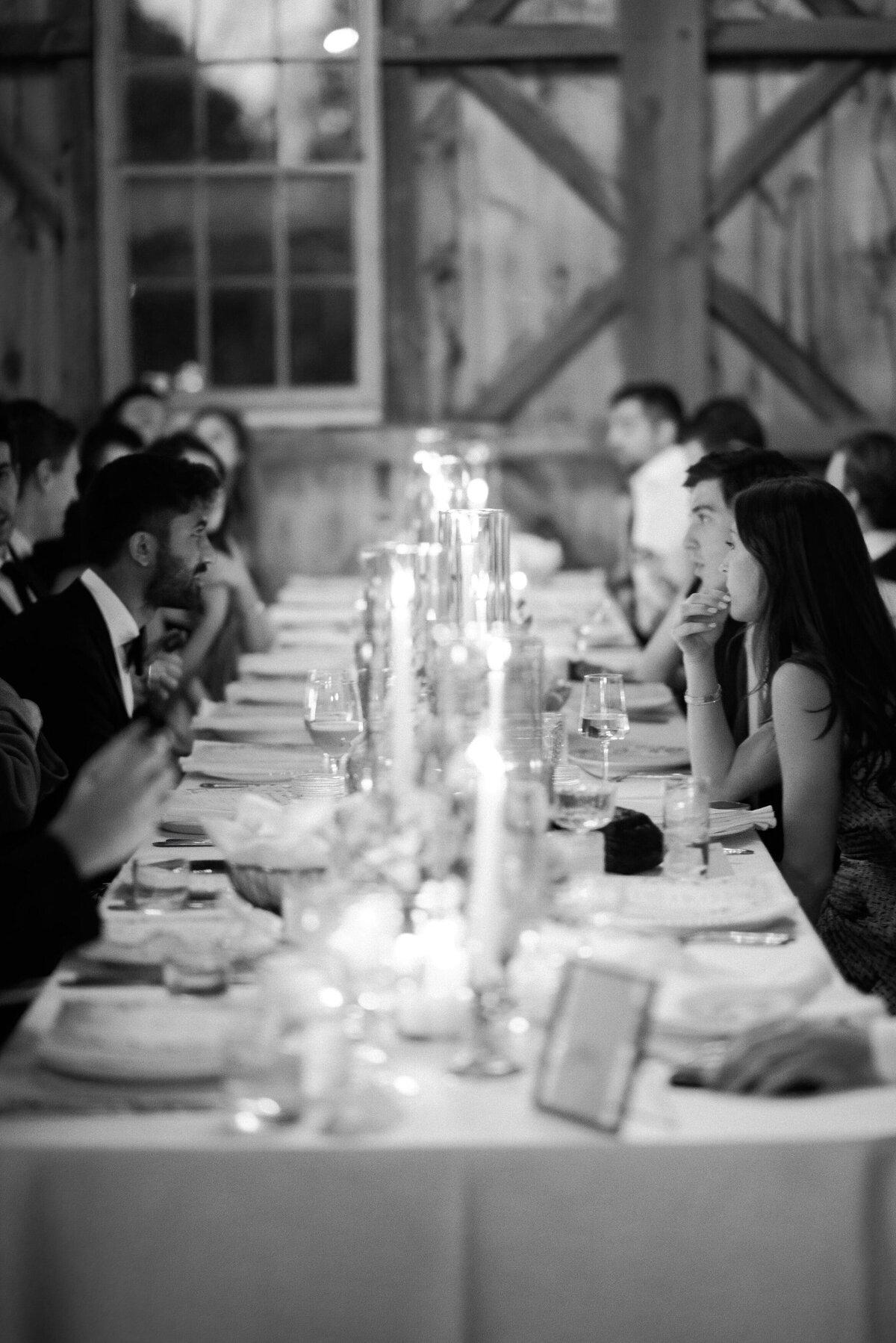 A long, elegant dinner table lined with flickering candles, surrounded by wedding guests inside a rustic barn.