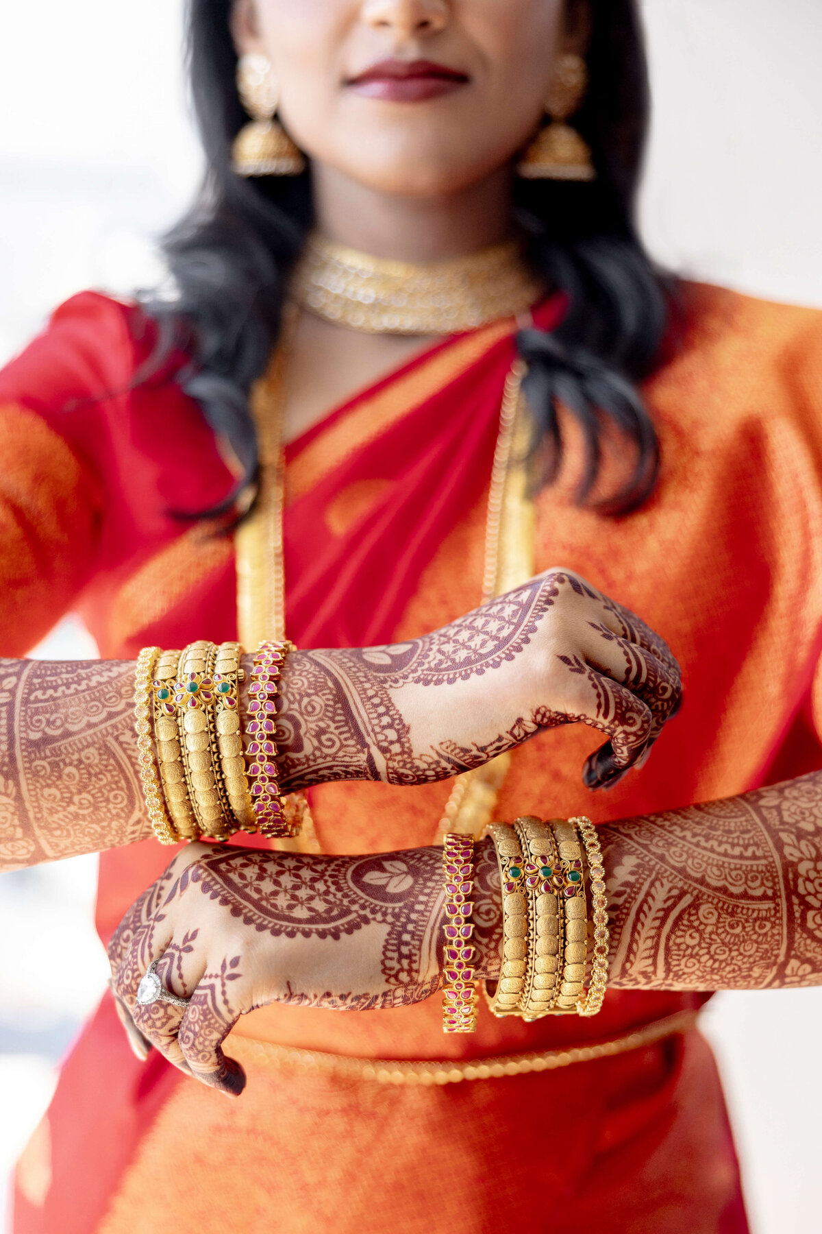A woman dressed in a vibrant red and orange sari showcases her intricately henna-decorated hands and gold bangles. She wears traditional gold jewelry around her neck and ears, adding to her festive attire.