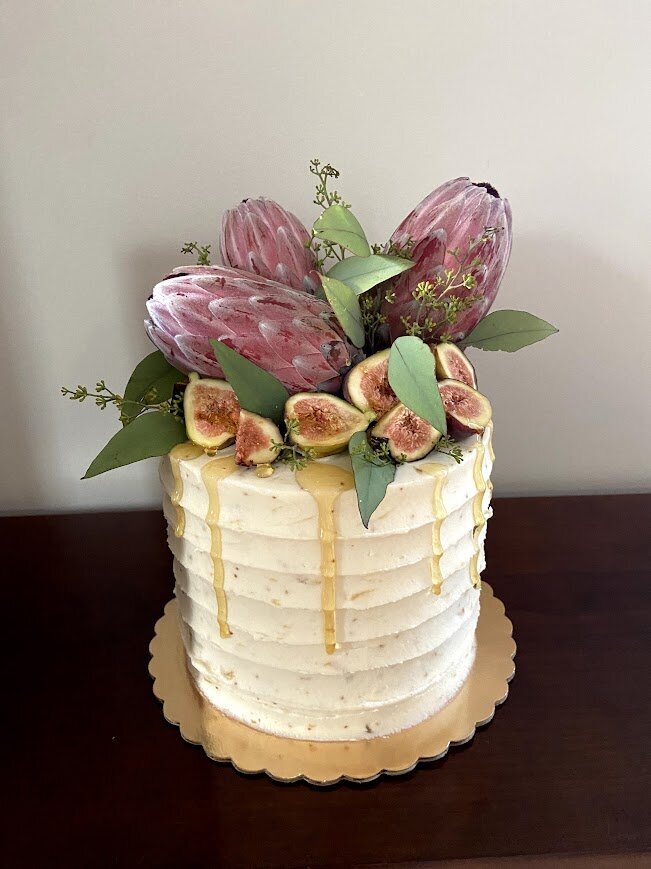 A small cake is decorated with fresh figs and protea flowers, and is dripping with honey.
