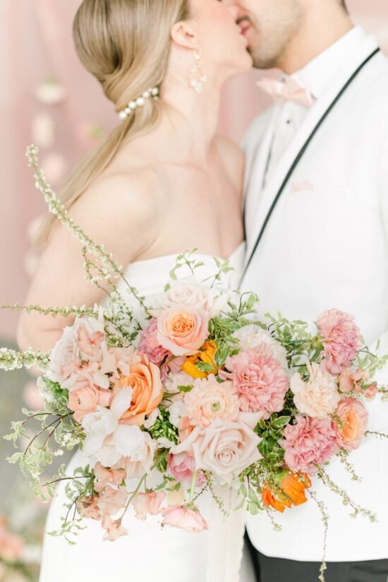 A bride and groom in white attire share a kiss, with the bride holding a large bouquet of pink and orange flowers, expertly arranged by their full wedding planner.