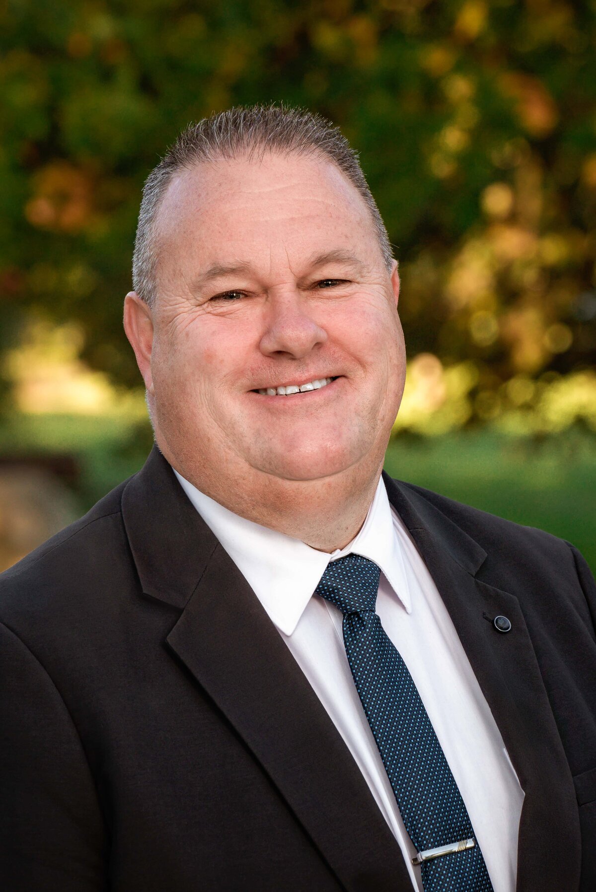 outdoor headshot of male real estate agent wearing white shirt, blue tie and black suit