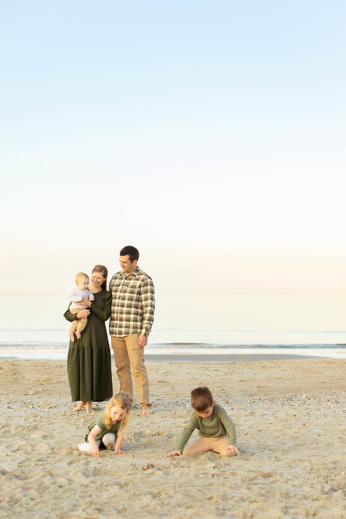 A young family enjoys a peaceful evening on the beach in Coastal Virginia, with mom and dad smiling at each other while their children explore the sand nearby.