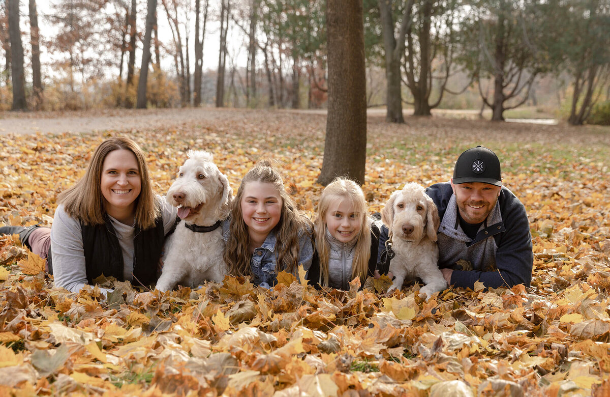 Family of 4 and their 2 large white dogs laying on their stomachs in a pile of leaves on a fall afternoon.