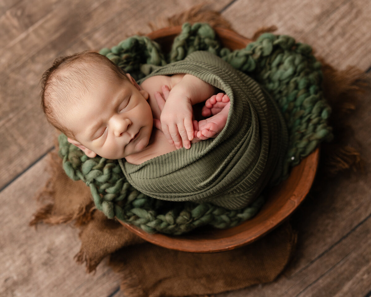 Newborn baby boy swaddled in deep green knits, posed in a wooden bowl with chunky textures and earthy tones. Captured by Inland Empire newborn photographer Kaitlyn Dawn Photography.