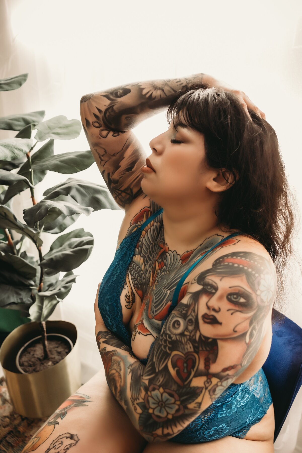 Tattooed woman sits on blue chair with her right arm up on her head with her head back and eyes closed