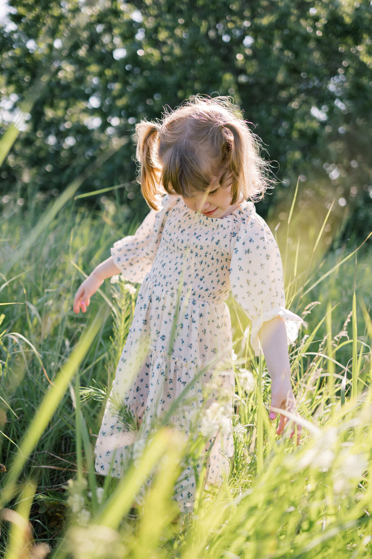 a young girl plays in a field of green tall grass as she smiles down at her hands, grazing the grass. She wears a white and blue floral dress and pigtails in Minnesota