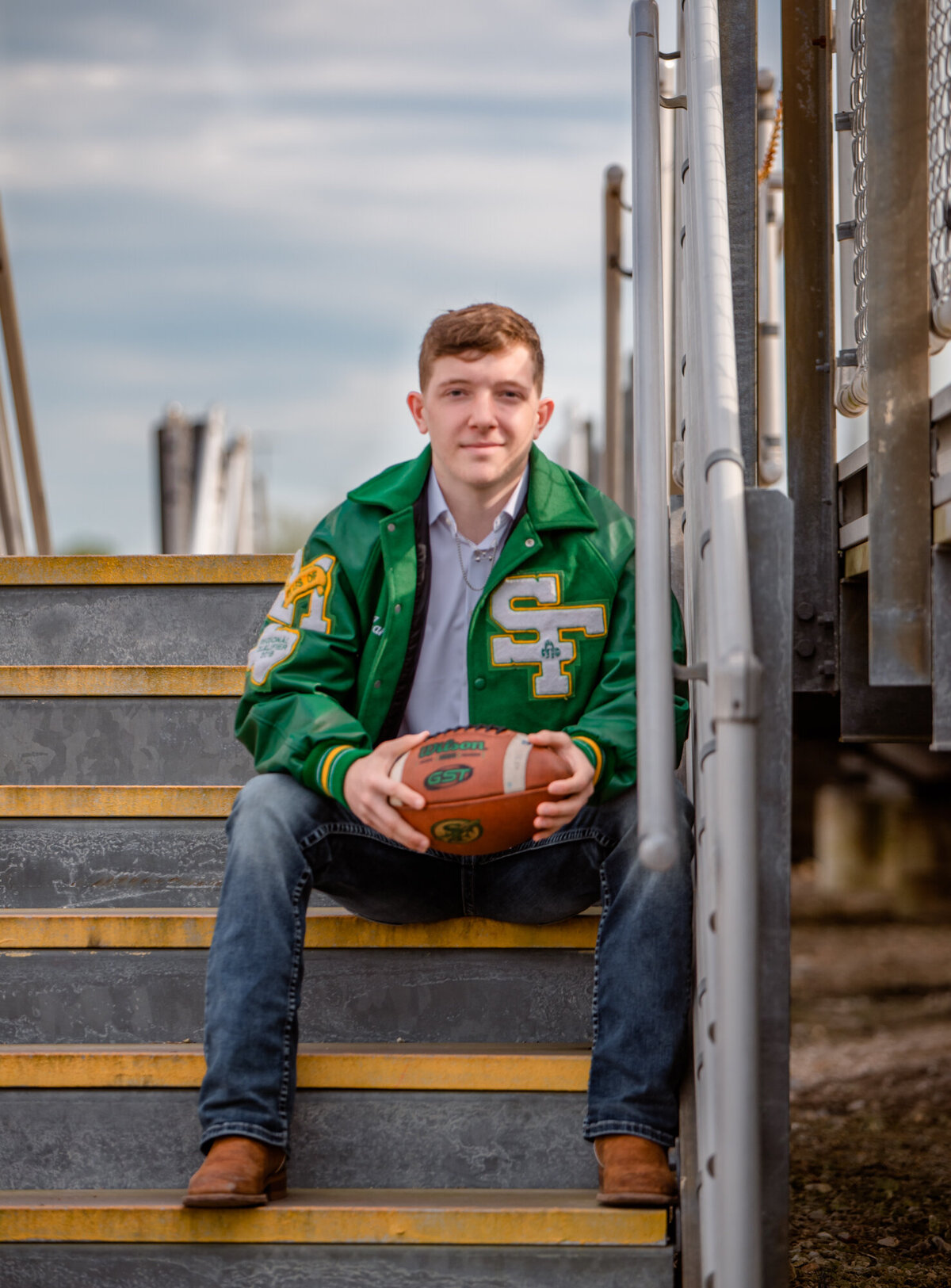 A senior wearing a letterman sits on the steps of football bleachers holding a football.