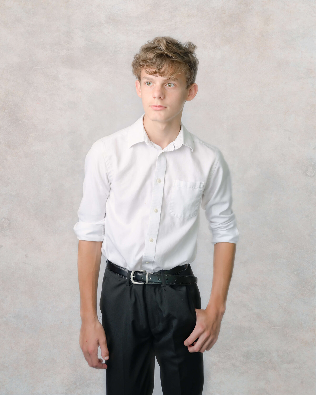 A teen boy dressed in white dress shirt and black pants looks off in the distance during his in-home studio-like  family photo session.