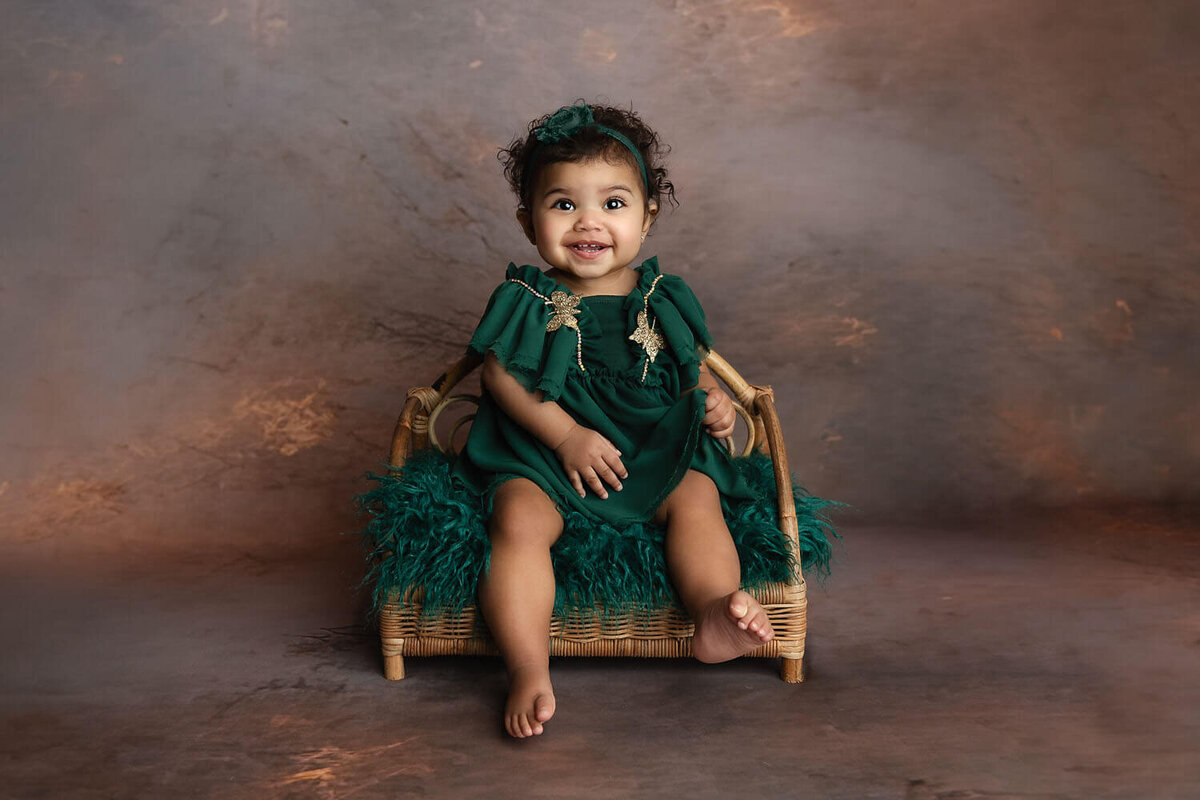 A toddler girl in a green dress sits on a wicker chair in a studio