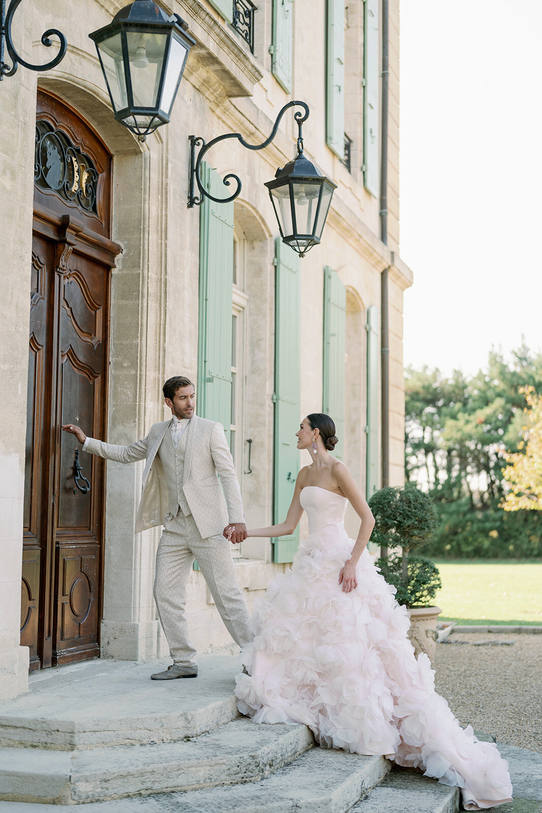 Groom opening ornate wooden door while helping bride step out of grand stone entrance at Château de Tourreau.