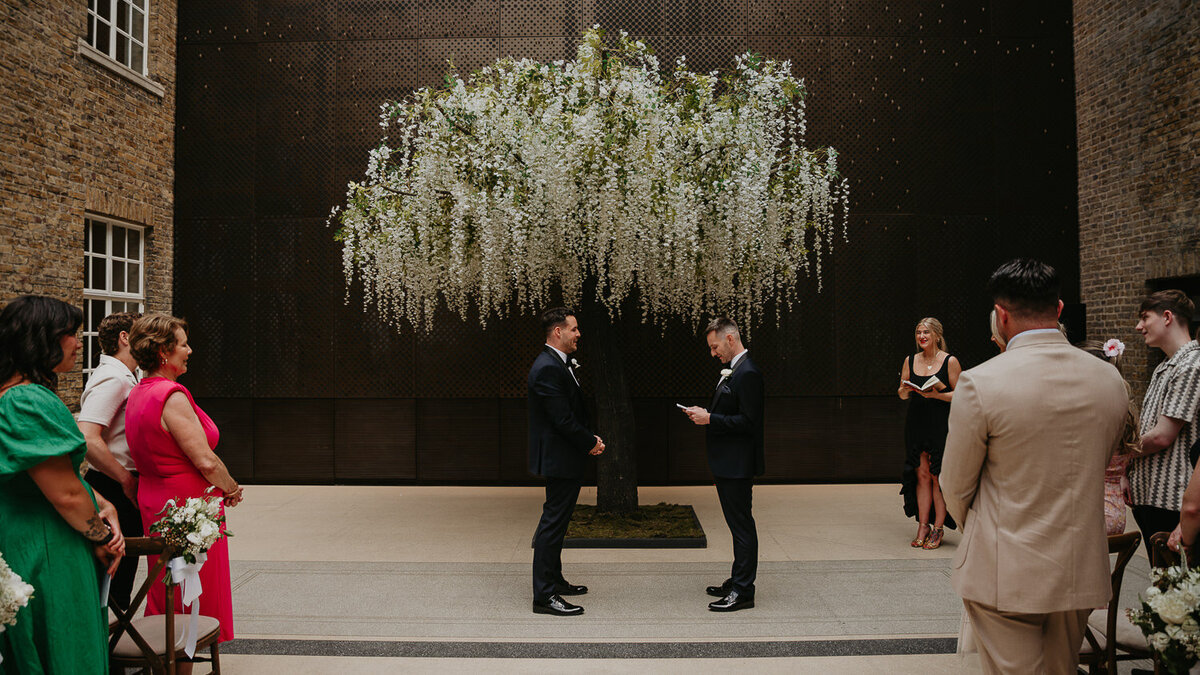 Two grooms get married at Hackney Town Hall in front of a tree. 
