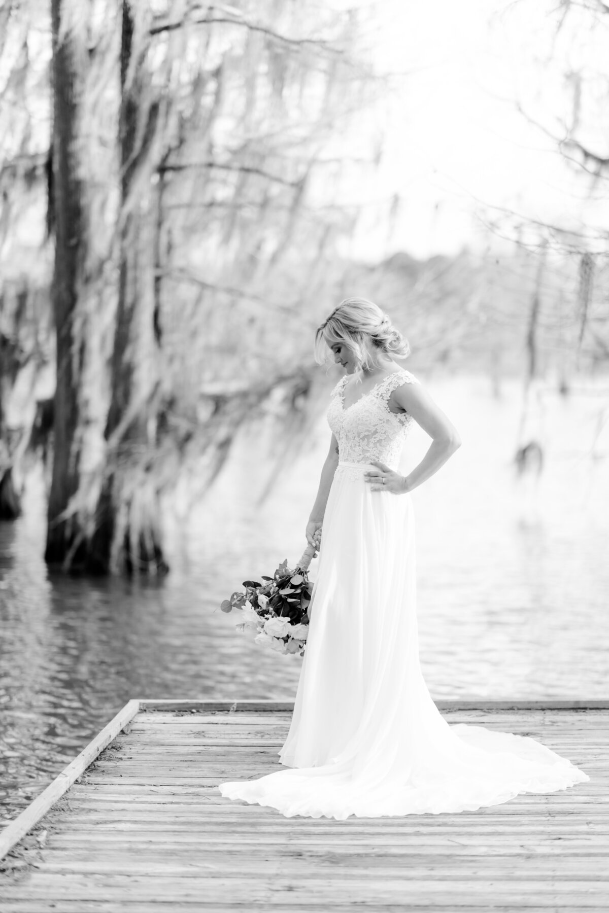 bride standing at the end of a dock with mossy hanging on the branches of trees around her as she looks down at her bridal bouquet