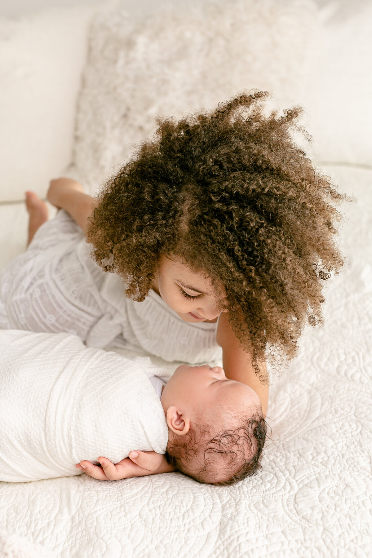 Sibling photo of older sister and newborn baby. Sister is about 4 years old and has dark curly hair, she is looking down at her brother and he is wrapped up in a white swaddle and looking back up at her. Image taken during newborn photography session in SE Portland.