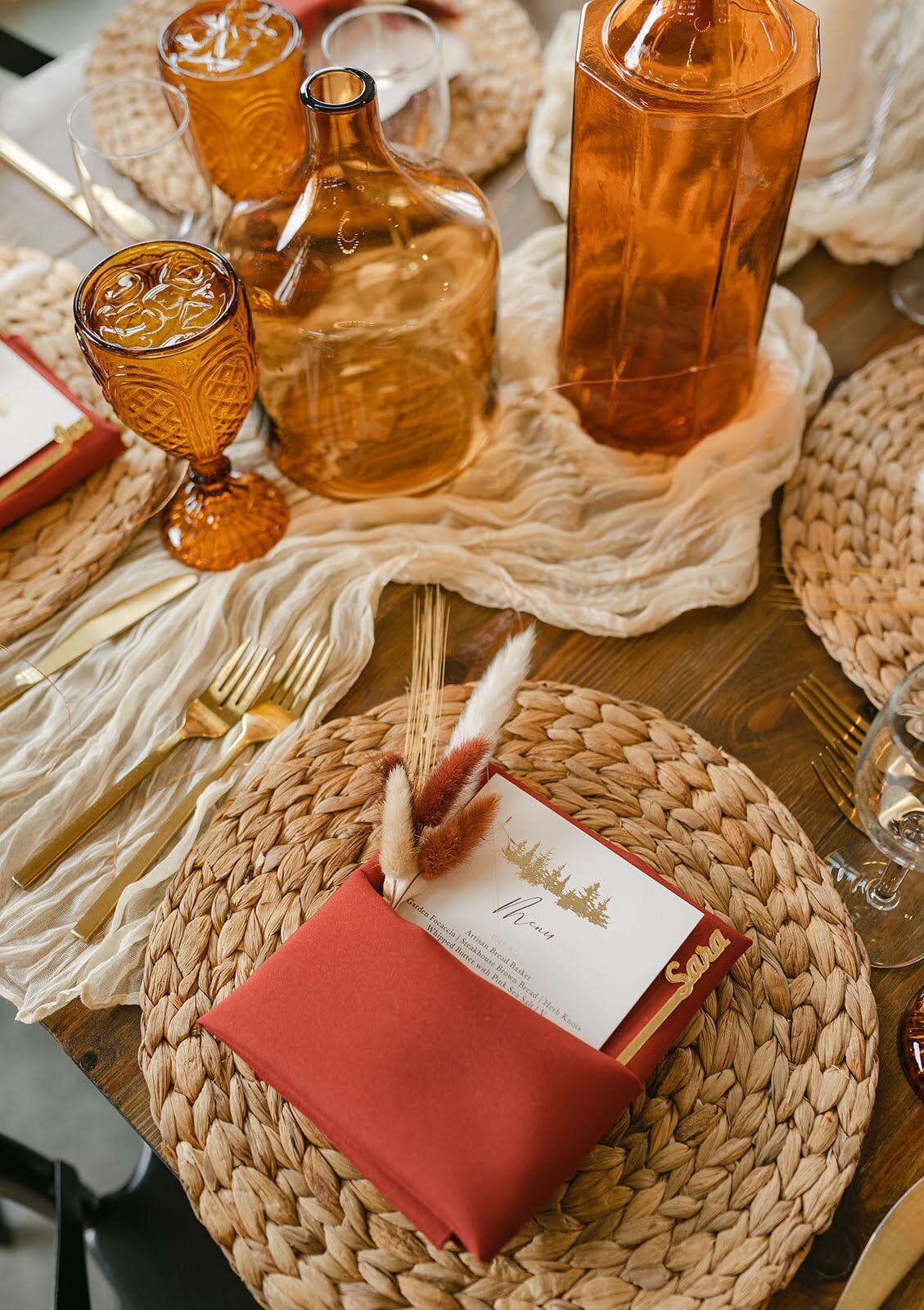 Wedding table place setting setup with warm colored vases, orange vases, wicker placemats, and gold flatware.