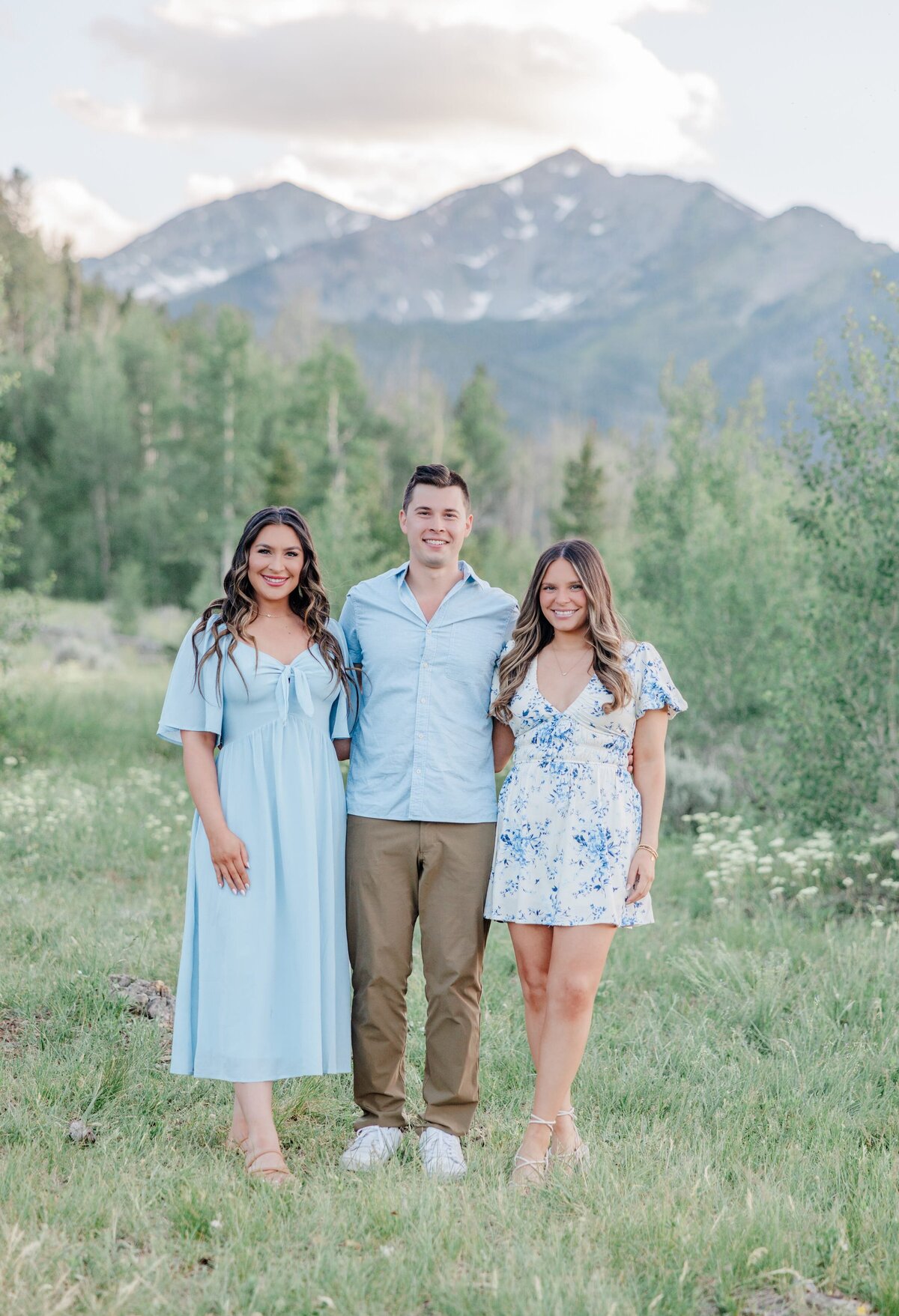 A man in a blue shirt stands with arms around his two adult sisters in blue dresses