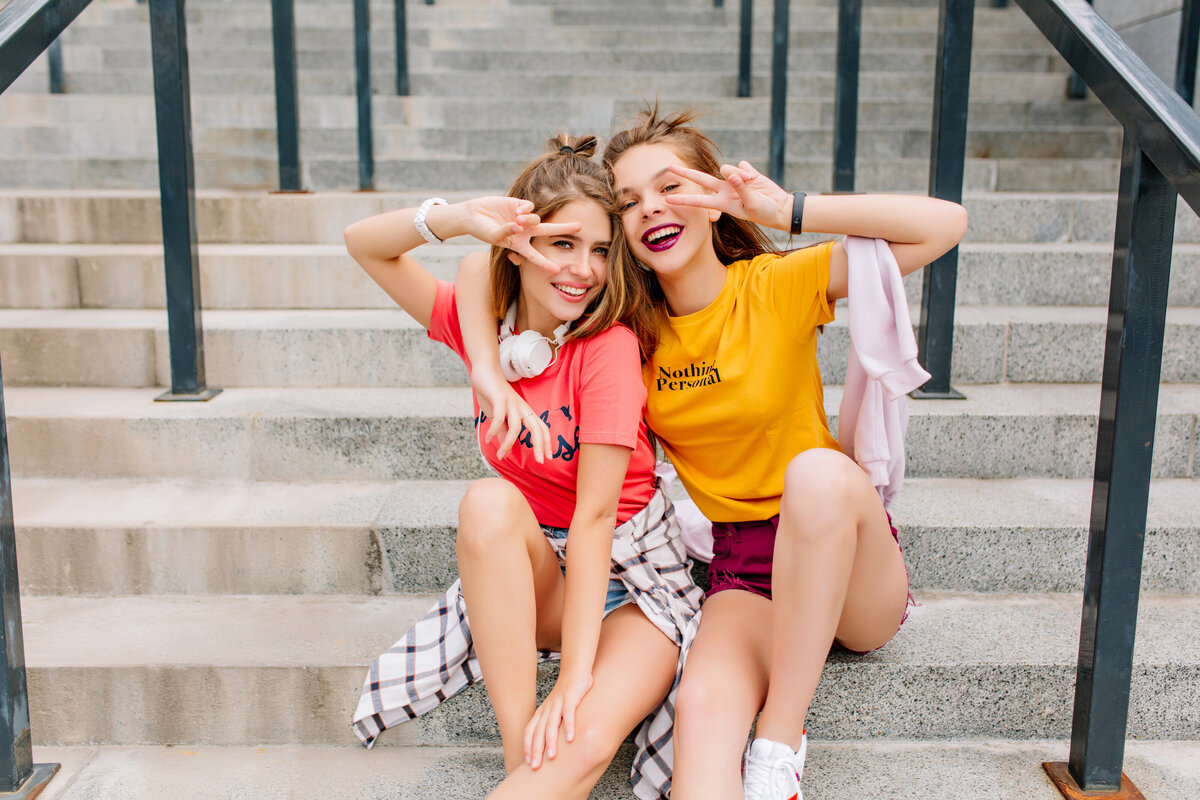 funny-inspired-young-ladies-trendy-outfit-gladly-posing-with-peace-sign-relaxing-stone-steps-summer-day
