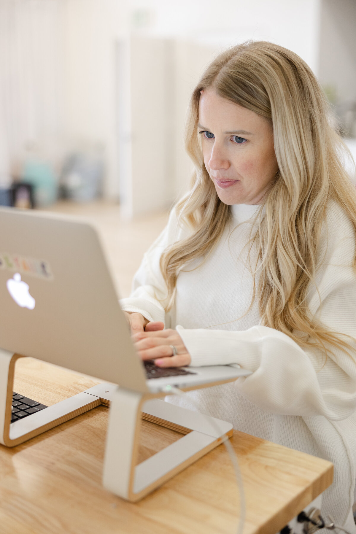 Woman with long blonde hair working on a laptop and phone in a bright studio office. Photograph by Yucaipa branding photographer Kaitlyn Dawn Photography.
