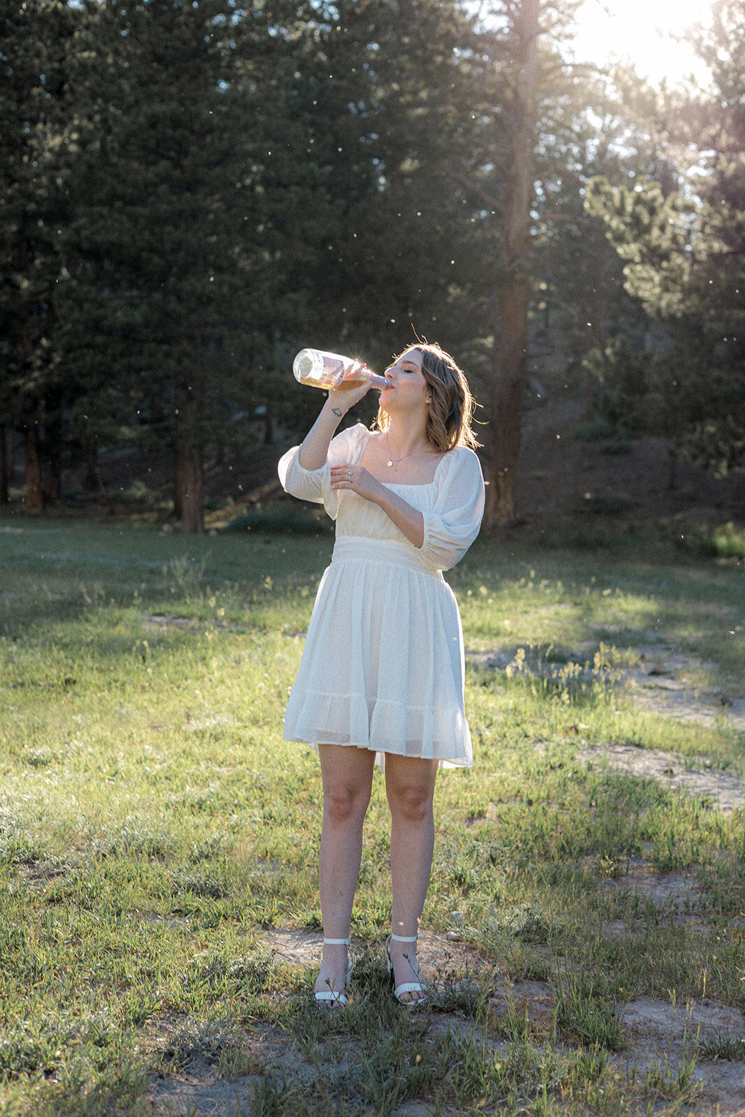 A woman in a flowy white dress drinks from a clear bottle, standing in a sunlit field with sparkles of dust catching the light around her.