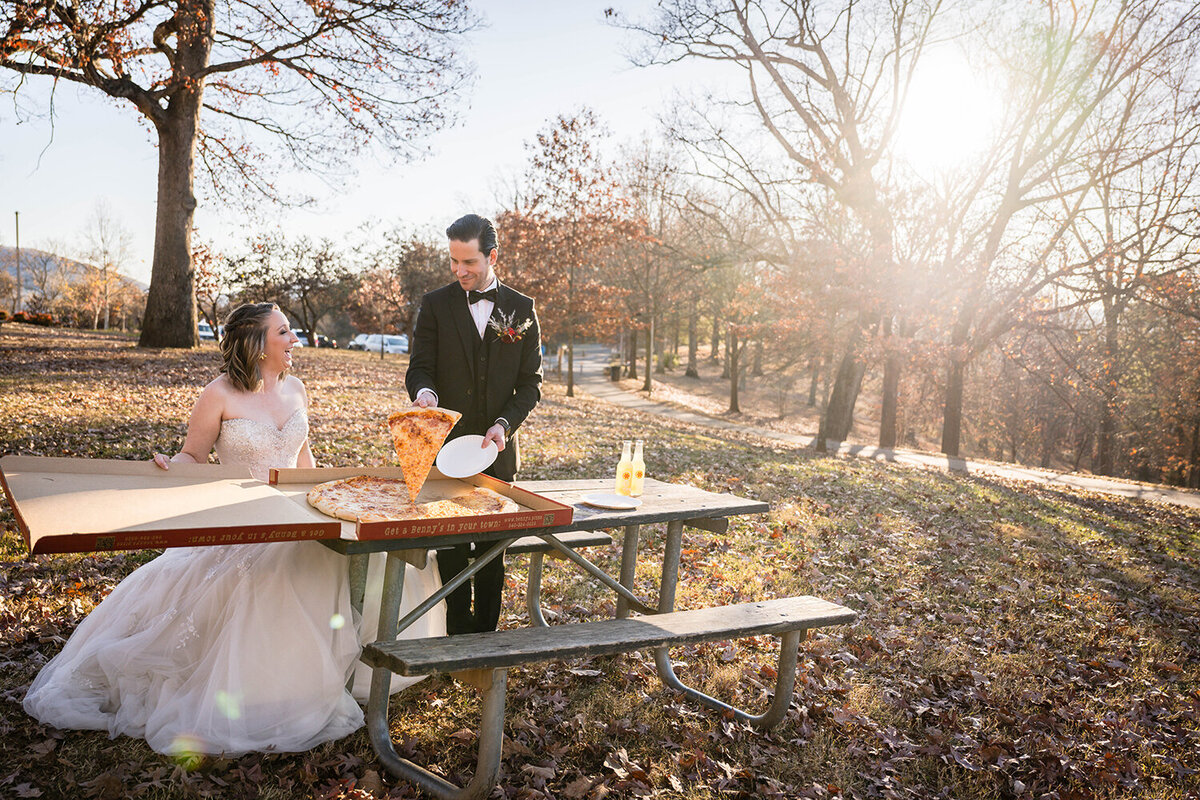 A groom stands and serves his significant other a slice of pizza at a picnic table near the Rockledge Overlook at Mill Mountain while the bride holds open the large pizza box and smiles up at her partner.