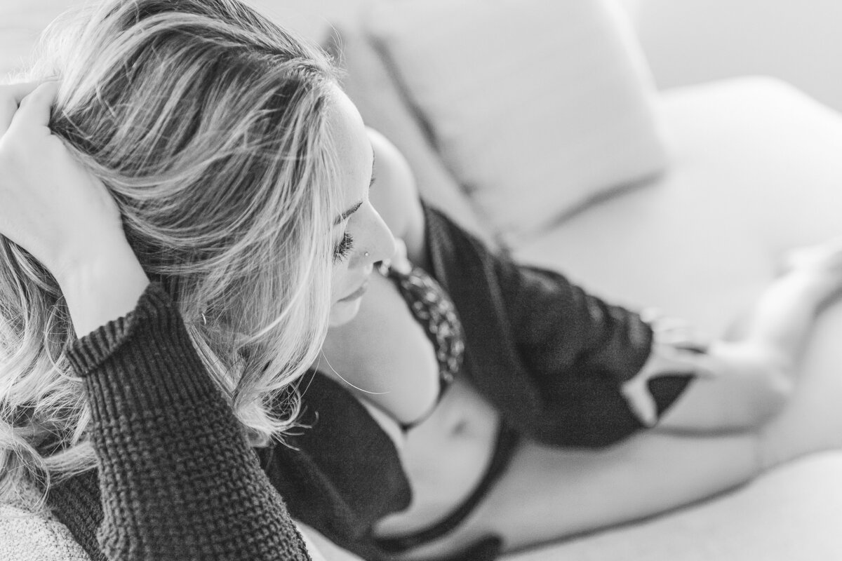 A woman lounging on a couch, wearing black lingerie and a cozy black sweater, gently running her fingers through her hair in a reflective mood.