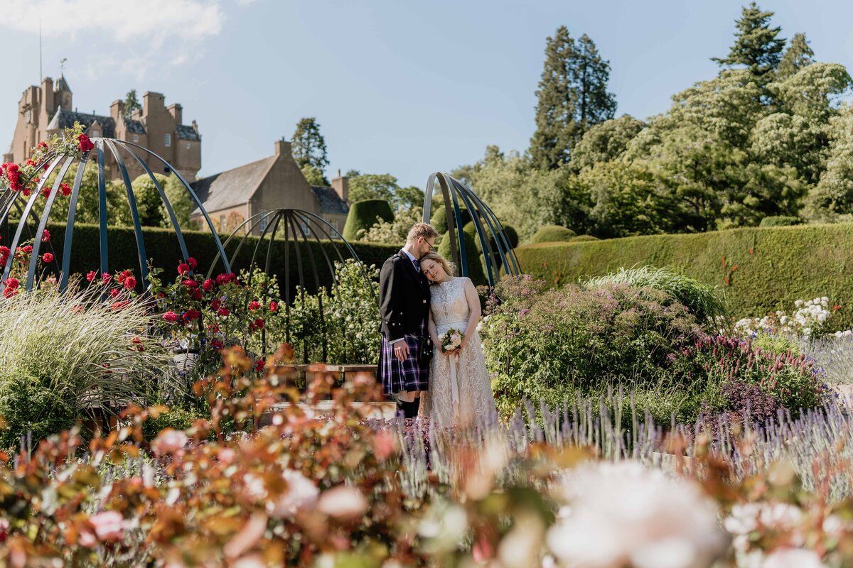 A newly married couple stand side by side in a large walled rose garden. They pose gently for photography and Crathes Castle stands in the background.