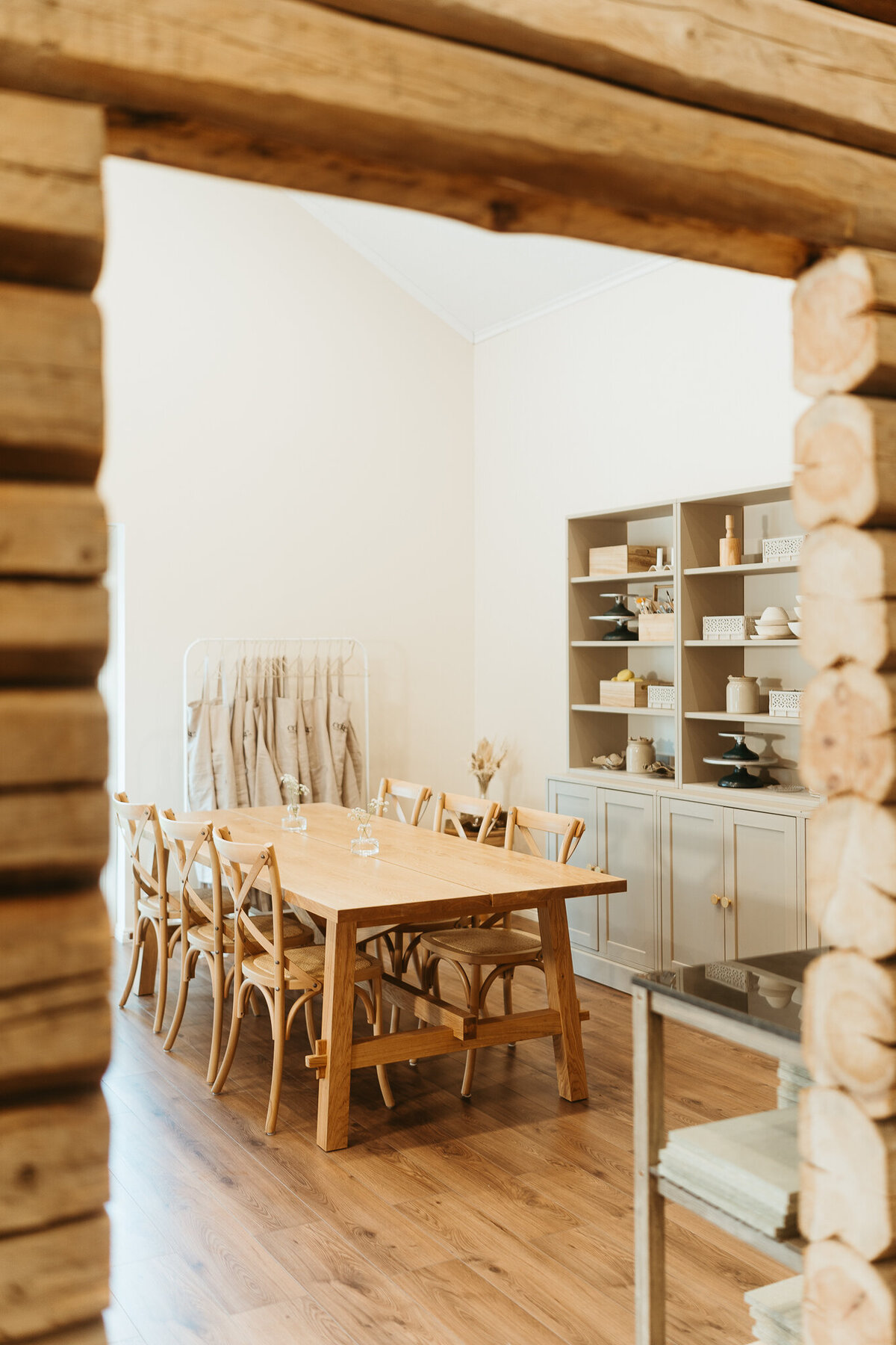 Kuvassa on keramiikkastudion tila, jossa näkyy kaksi pöytää ja niiden tuolit, essuja naulakossa ja perällä hylly täynnä erilaisia työvälineitä.