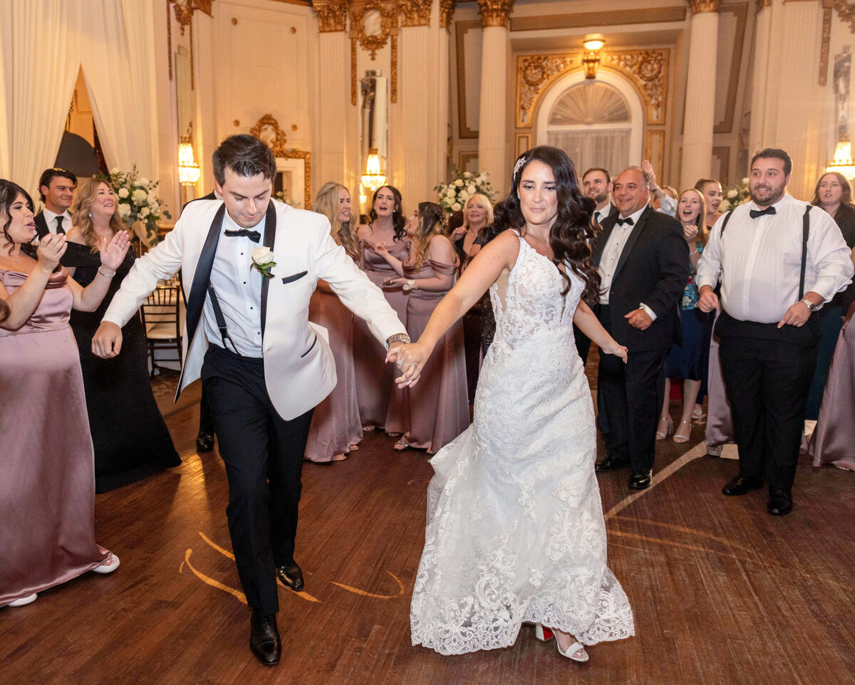 A bride and groom dance energetically at a wedding reception. The bride wears a white lace dress and the groom a white suit jacket with black pants. Guests, dressed formally, cheer and clap around them in a grand ballroom with ornate decorations.