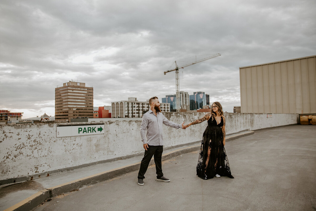 Engagement photoshoot at the top of a downtown London, Ontario parking garage. The man and woman are walking and holding hands. the woman is leading the man and looking over her shoulder at him.