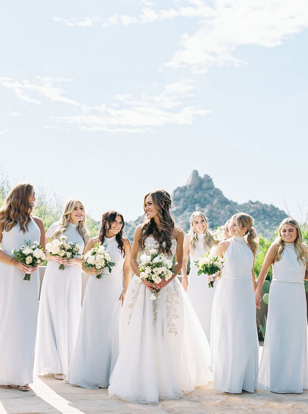 The bride and her bridesmaids pose in elegant white gowns against a stunning Arizona desert backdrop, showcasing timeless wedding fashion.