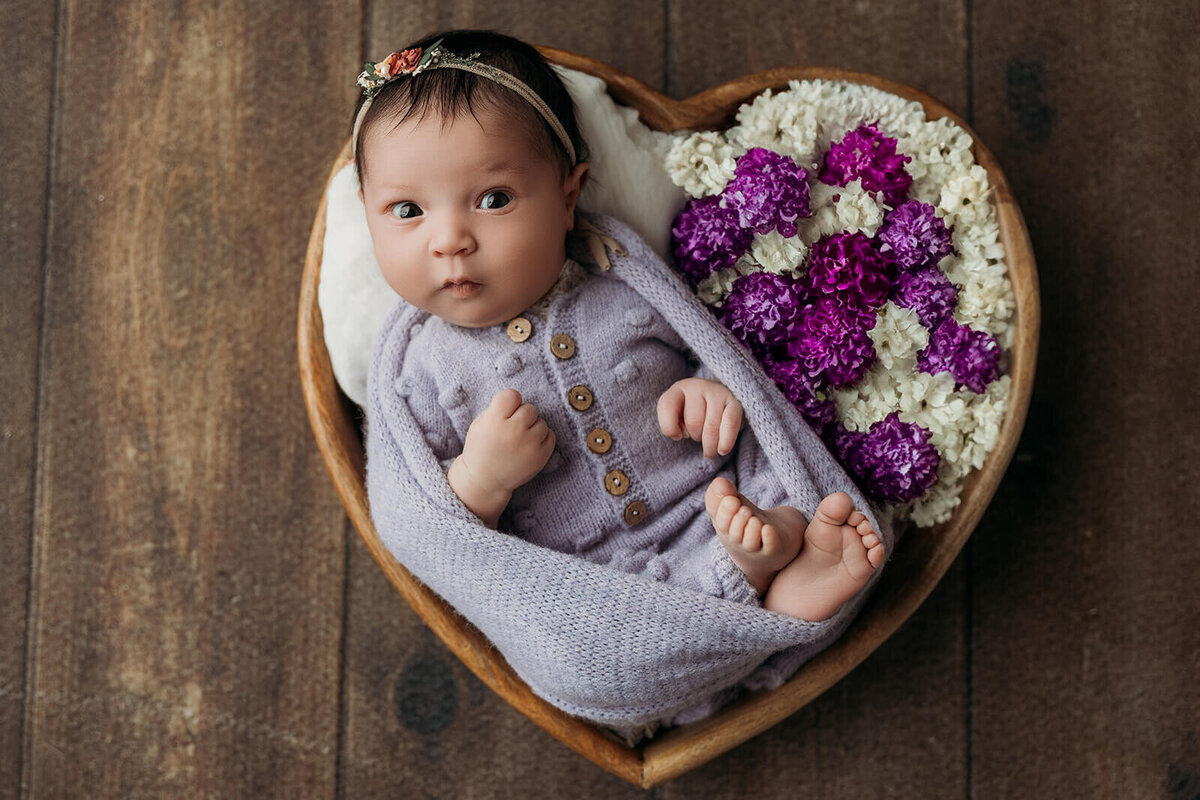 A newborn baby, wearing a light purple outfit, rests peacefully in a heart-shaped basket filled with white and purple flowers. The baby looks up curiously, with a small headband adorned with a flower. The rustic wooden background adds warmth to the scene.