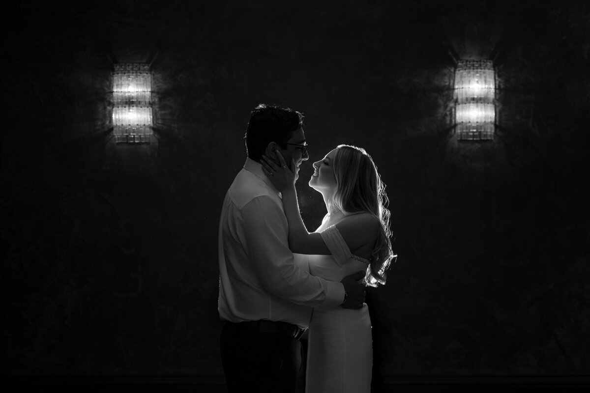 Black and white image of a couple sharing an intimate moment, lit dramatically from behind to highlight their profiles and soft movement in the hair, flanked by glowing wall sconces—an example of moody editorial NYC engagement photos that blends cinematic lighting with a quiet, emotional connection.