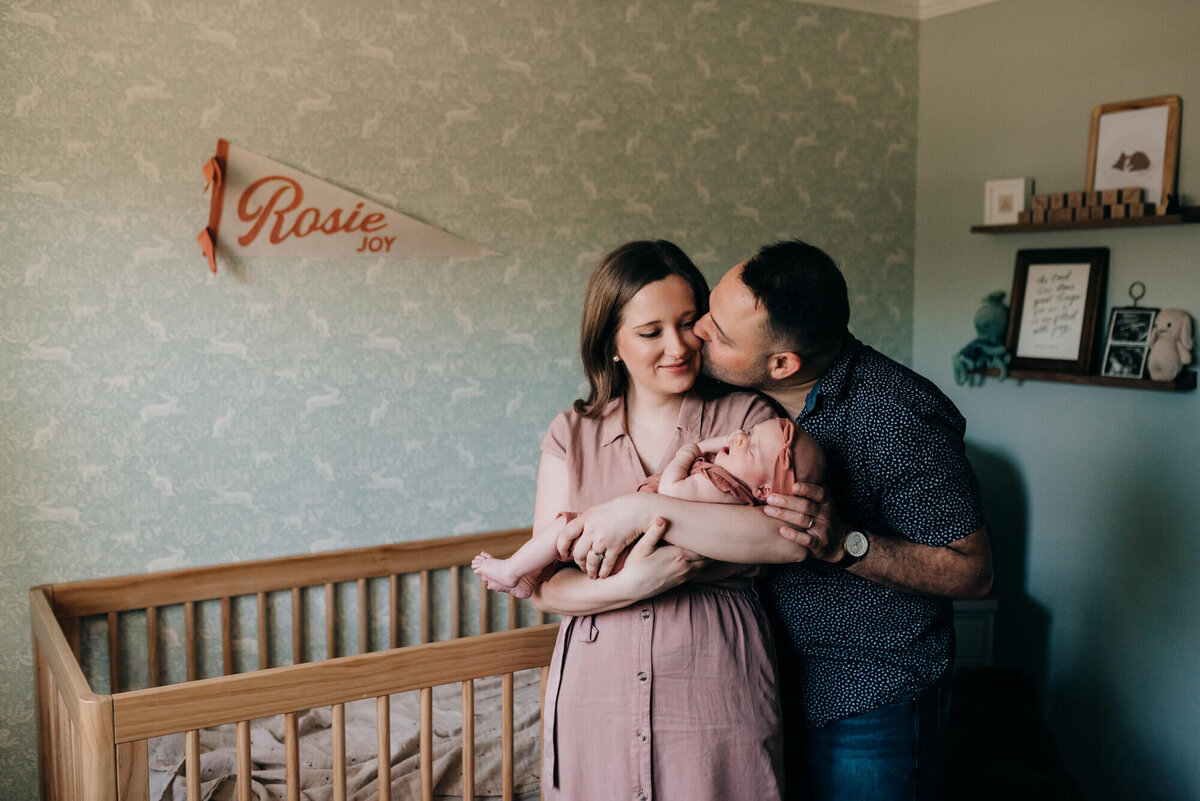 A mother and father hold their newborn daughter in her nursery, the father kisses the mother's cheek during their photoshoot with Boston Newborn Photographer, Allison Wolf