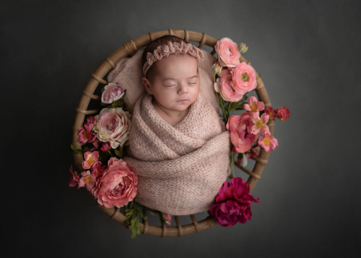 baby wrapped in pink with pink headband in a bowl with pink flowers and a dark gray background