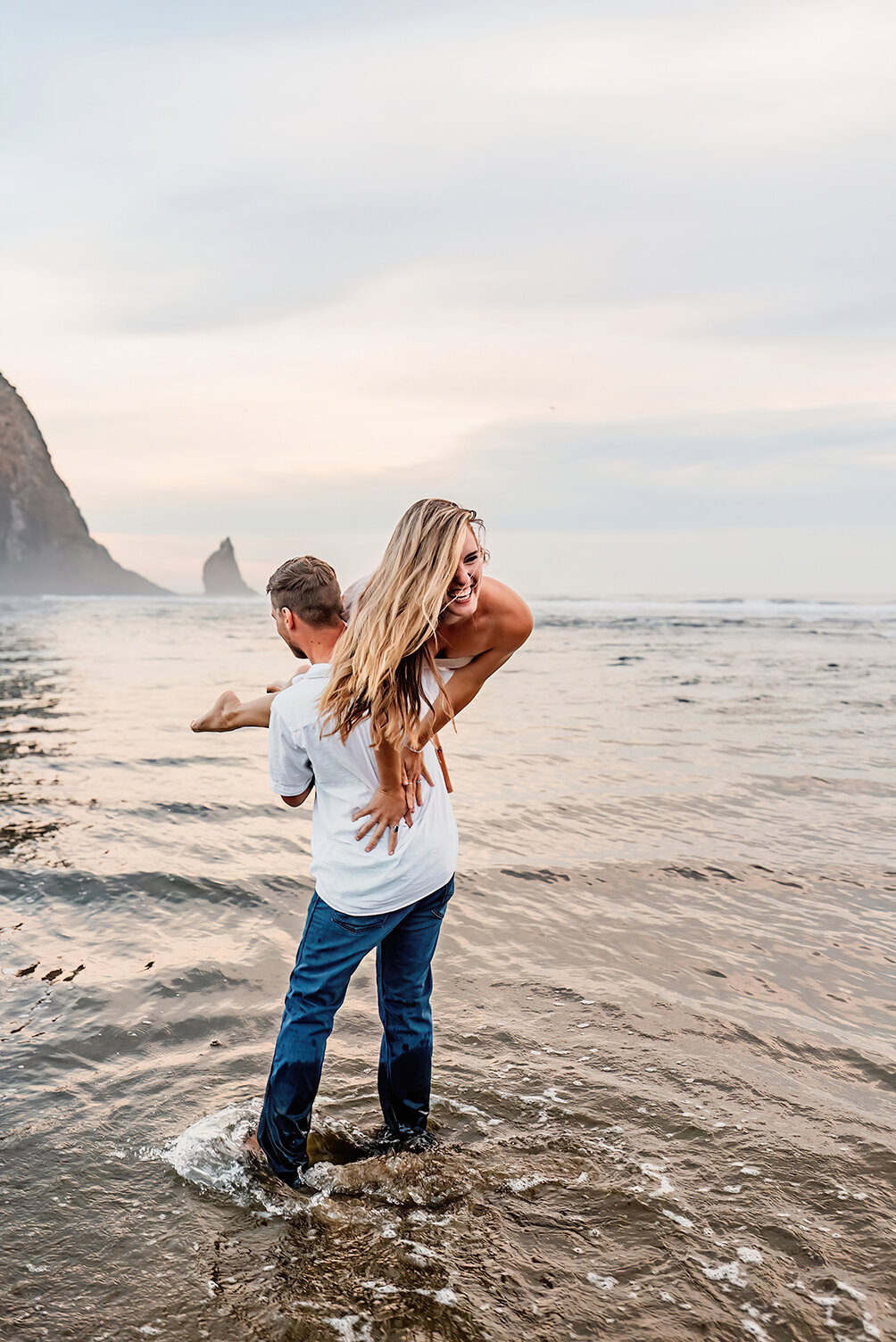 a women over her husbands shoulders laughing on the beach next to Haystack rock, Cannon Beach, OR