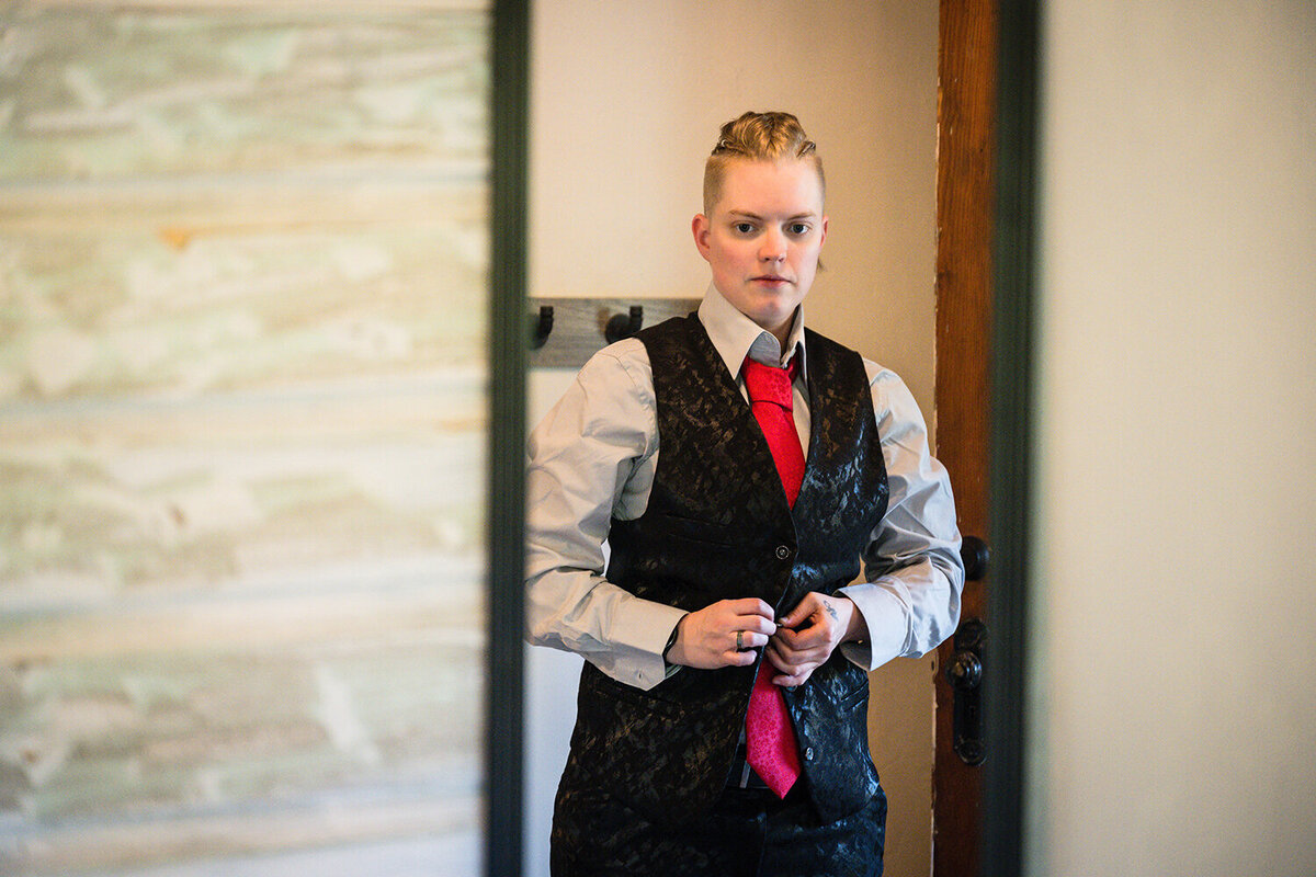 An LGBTQ+ bride buttons her suit vest while looking in a mirror in another room in the Airbnb on her elopement day.
