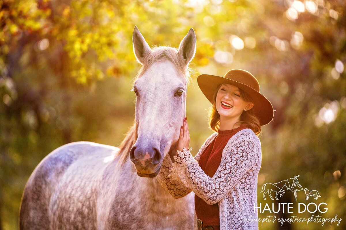 Female equestrian wearing a felt hat smiles as she holds her gray mustang's face under a soft glow.