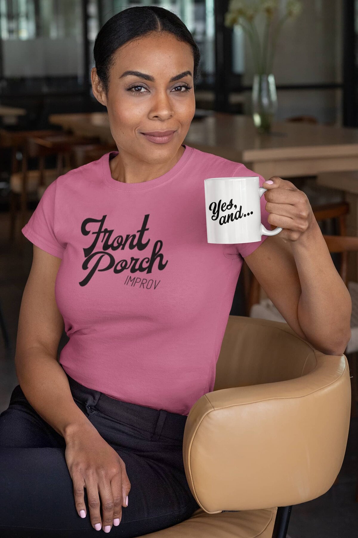 A Black woman sits in an office chair wearing a pink Front Porch Improv t-shirt and holding a white coffee mug with the words "Yes, and..."