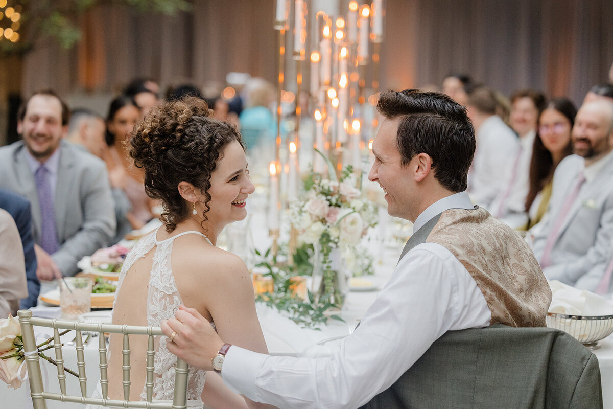Bride and groom looking at each other at reception table with florals and candles in the background