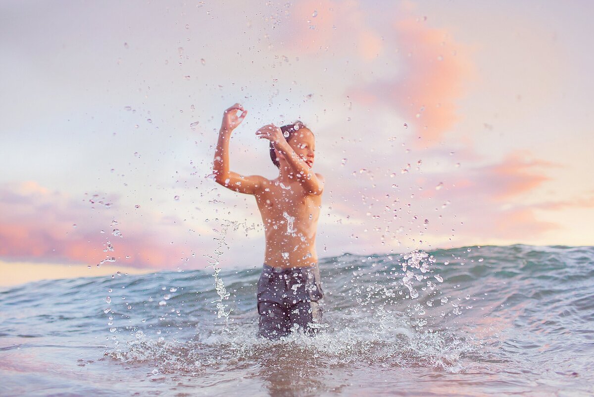 Boy in swim trunks splashes water in the air as waves break behind him in a beautiful family photo by Love + Water Photography