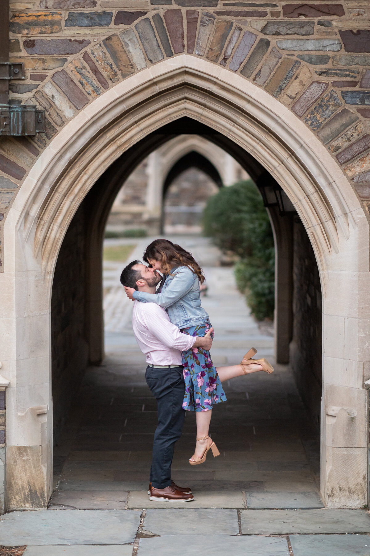 A man lifts and kisses a woman under a stone archway in an outdoor walkway with patterned stone walls in the background