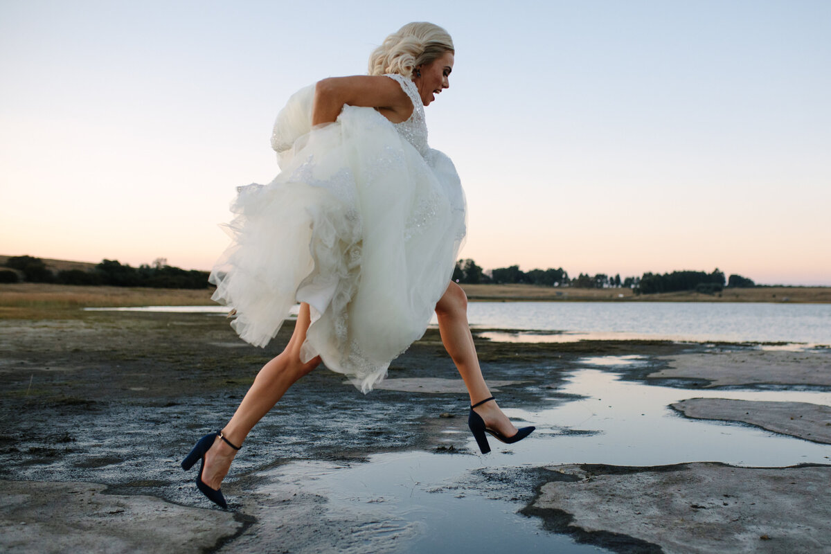 Spontaneous bridal jump over a puddle, dress held high and blue heels on display, captured in a joyful, natural moment.