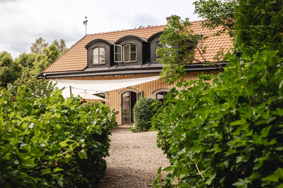 Charmigt gästhus med träfasad och tegeltak omgivet av grönska – ett lantligt pensionat i sommaridyll.