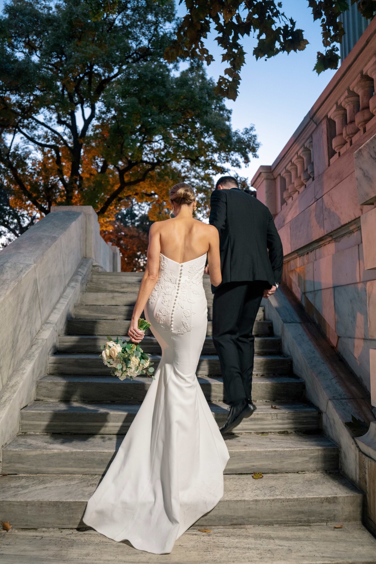 A bride and groom are walking up stone stairs. The bride is wearing a white strapless gown and holding a bouquet, while the groom wears a black suit. The scene is outdoors, with trees and a pastel-colored sky in the background.