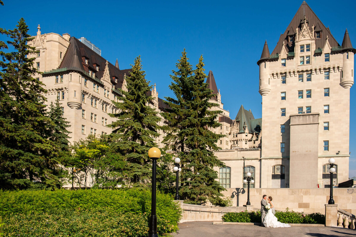 scenic photo of the Chateau Laurier wedding venue with a big blue sky and a bride and groom kissing in front.  Captured by Ottawa wedding photographer JEMMAN Photography