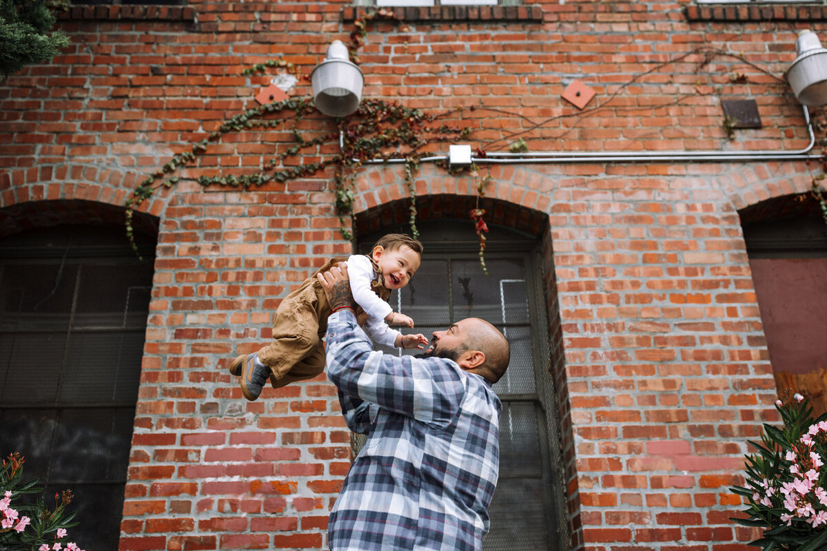 dad with tattoos and plaid shirt holding toddler son above his head as they both laugh in front of brick building in Oakland