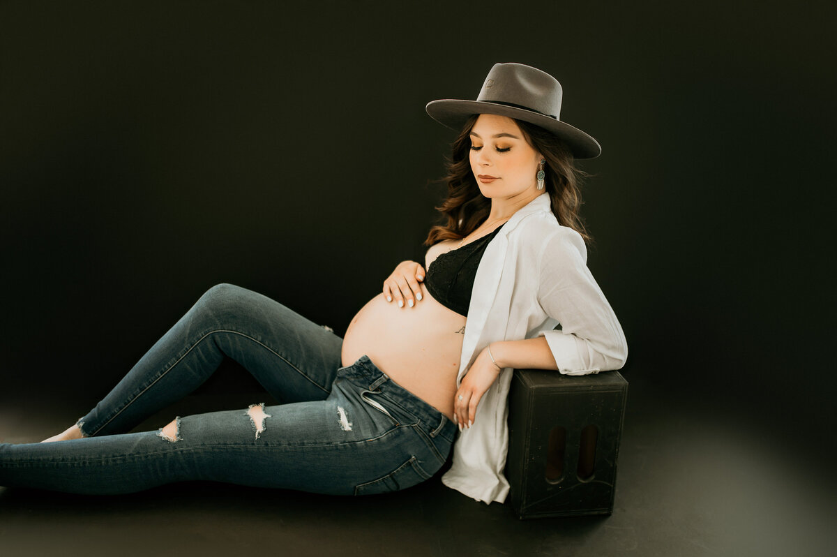 Pregnant woman reclining on a black stool wearing jeans, a black bralette, a white button-up, and a wide-brimmed hat, captured in a minimal, contemporary maternity portrait.
