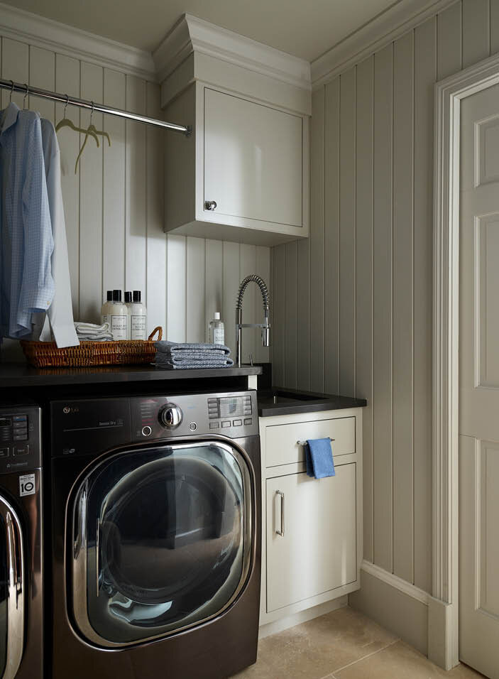 Discover the functional laundry room in a historic Hingham home, with custom cabinets, limestone flooring, light grey shiplap, ample counter space, and a hanging rod.