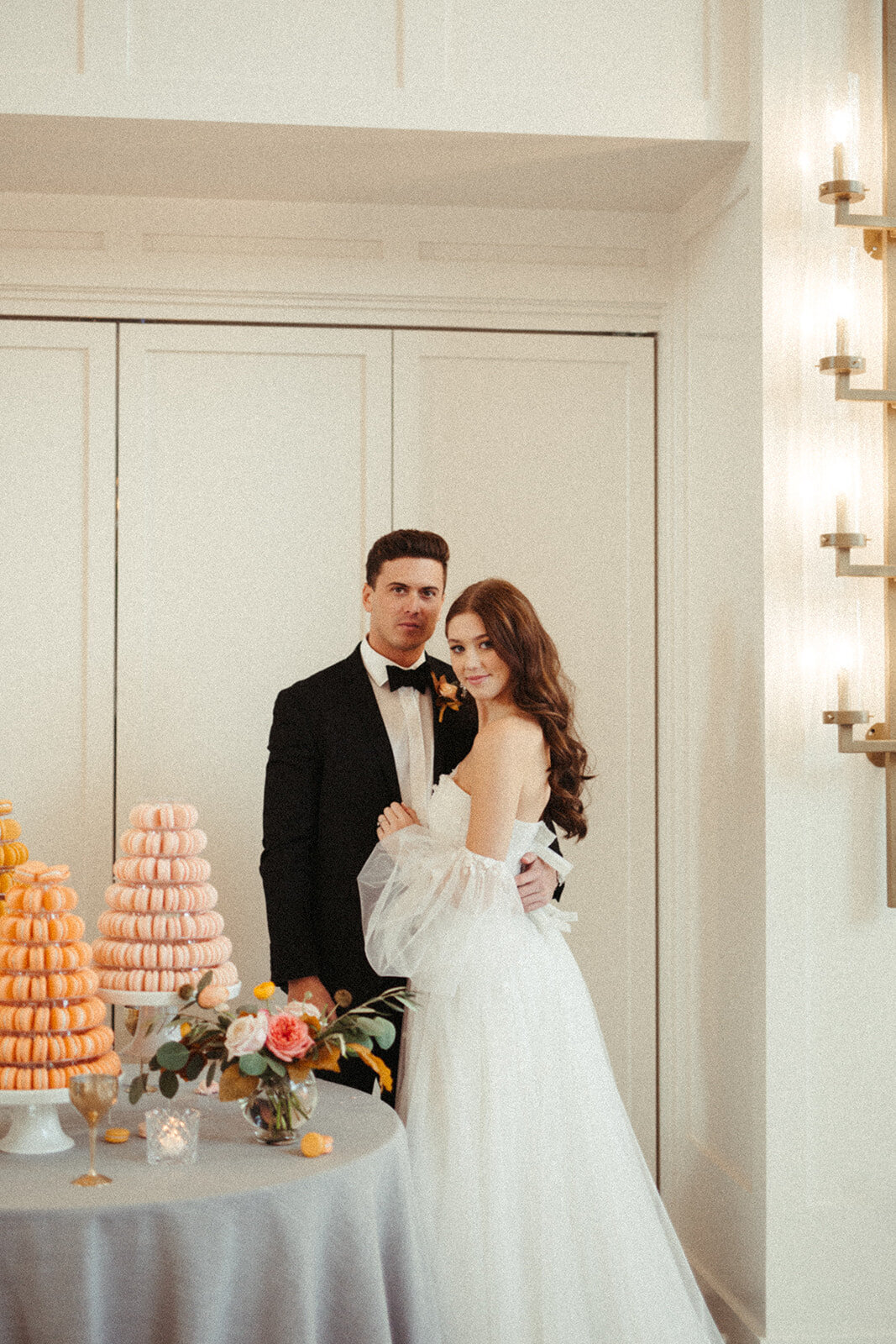 A bride and groom wearing a white wedding gown and black tuxedo lean stand next to a dessert table filled with macarons.